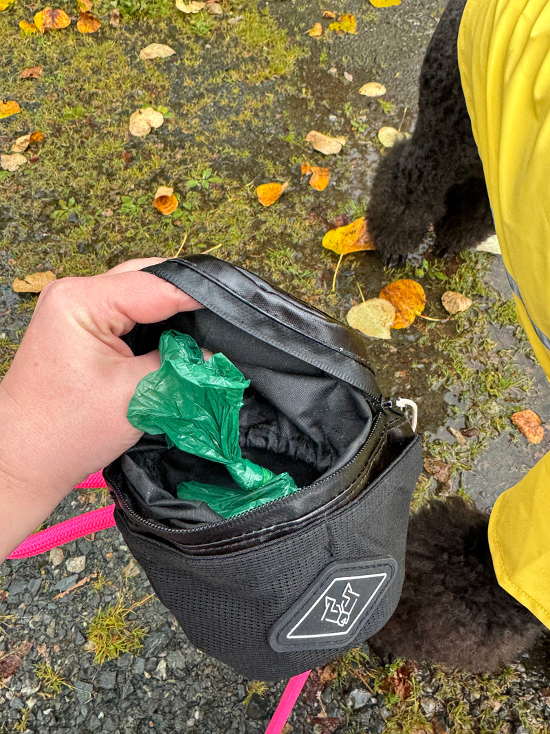 Green Eco Poop Bags roll inside an open Black Waste Wrangler on a rainy walk, a dog in a yellow raincoat beside it