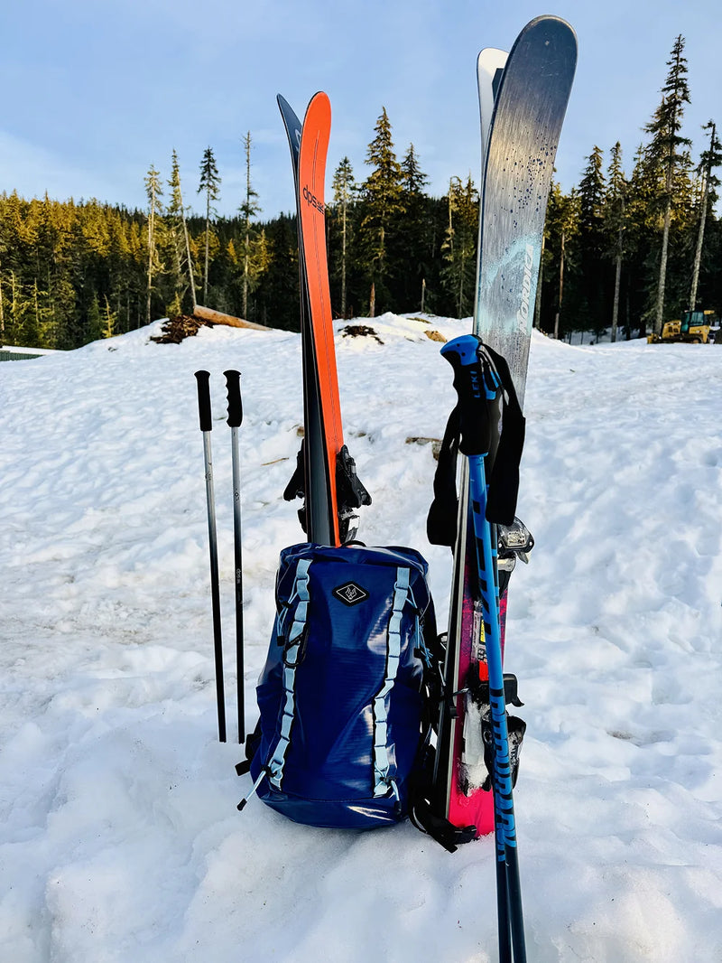 Blue Wolf Stride backpack positioned beside skis and trekking poles in a snowy mountain setting