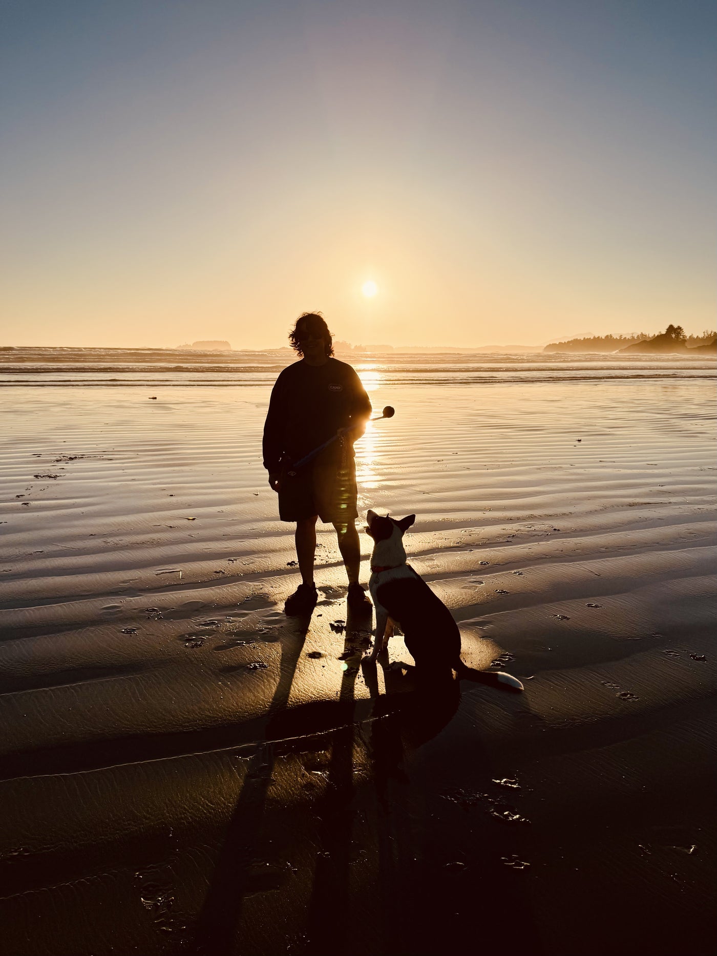 Jessie sitting on a Vancouver Island beach at sunset, looking up at Jennifer holding a ball launcher