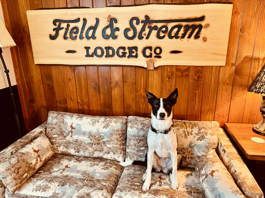 Jessie sitting upright on a fishing-print couch beneath the carved Field and Stream Lodge Co. sign