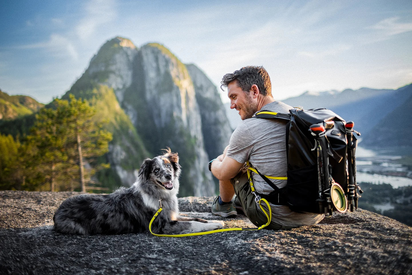 Smiling hiker with loaded adventure backpack sharing a quiet mountain summit moment with his happy border collie — built for life with your dog